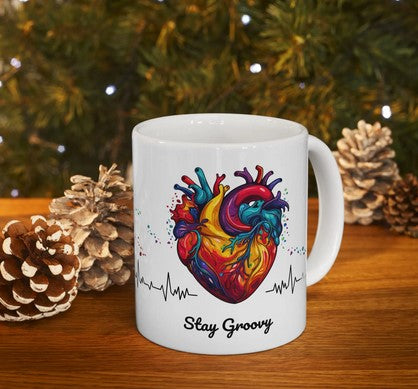 White mug with colorful heart design and 'Stay Groovy' text, placed on a wooden surface with pinecones and a Christmas tree in the background.